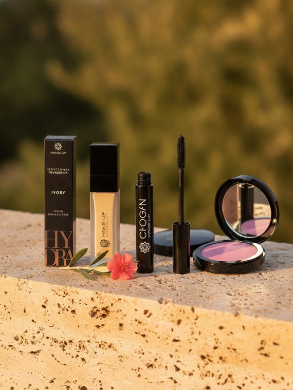 MoniAmori Make-up, Foundation, Mascara und Rouge, arrangiert auf einem Travertinblock vor einem Wasserfall in warmem Oasenlicht