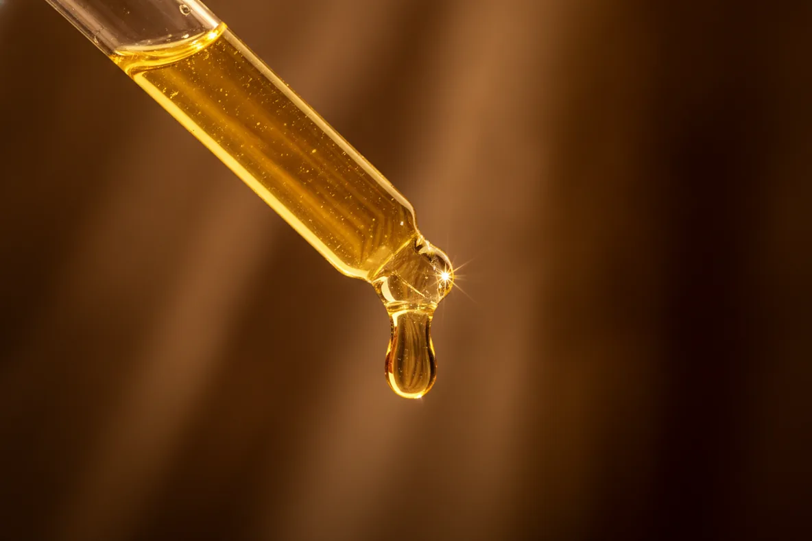 Makroaufnahme eines goldenen Serumtropfens an einer Glaspipette im Gegenlicht