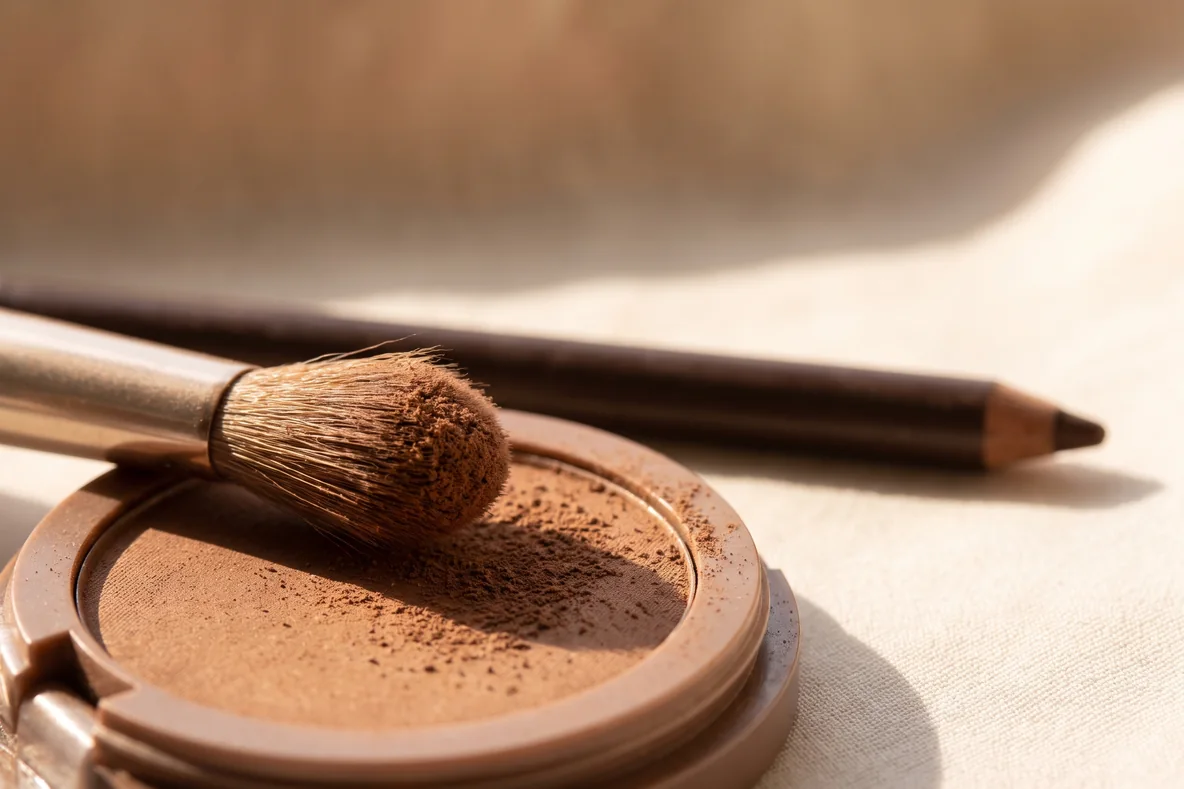 Ein matter brauner Lidschatten, ein Kajalstift und eine Mascara liegen nebeneinander auf hellem Leinen in warmem Licht