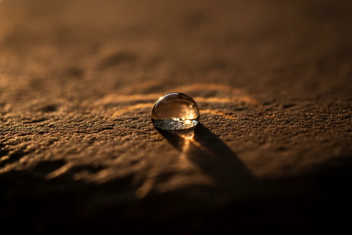Makroaufnahme eines Wassertropfens auf dunklem Stein im harten Sonnenlicht