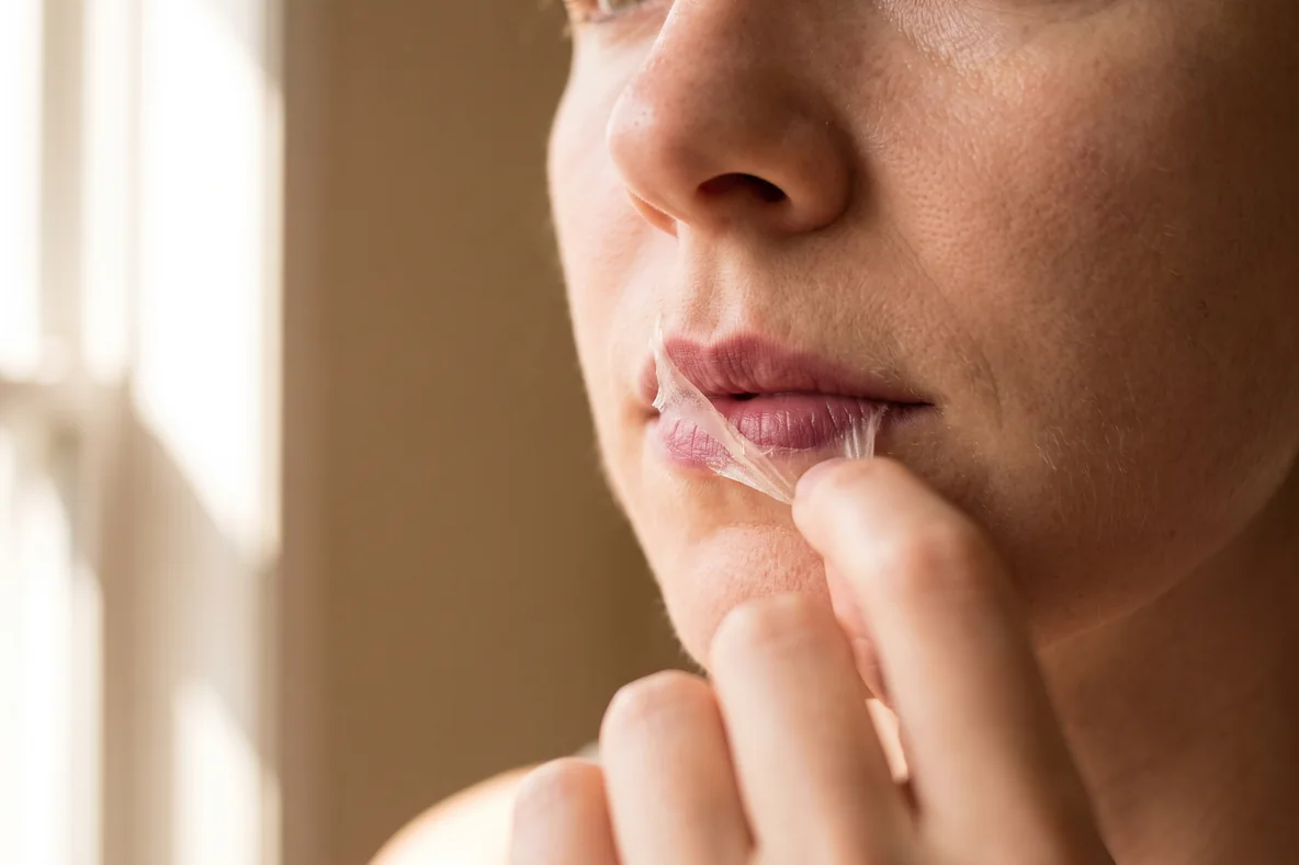 Nahaufnahme von Lippen, an denen ein getrockneter, transparenter Farbfilm vorsichtig von einer Ecke abgelöst wird, in warmem Licht