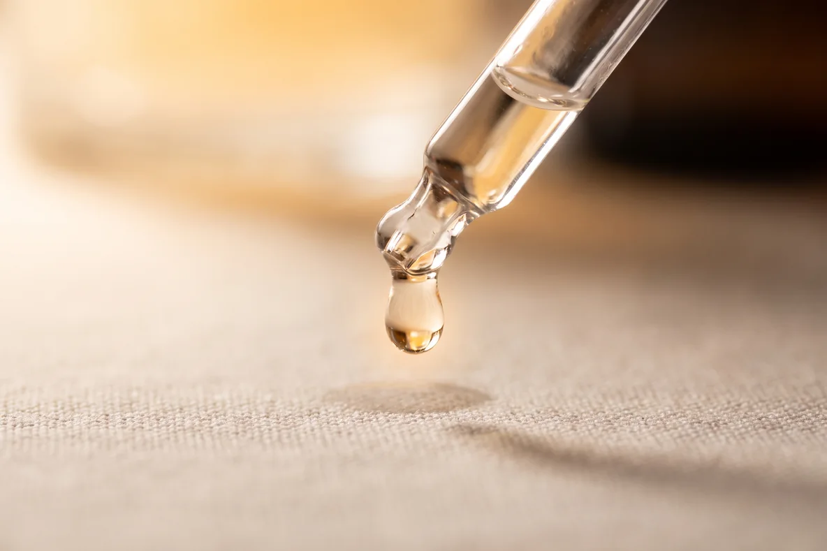 Eine Pipette mit klarem Serum schwebt über einer hellen Leinenfläche, ein Tropfen hängt an der Spitze, in warmem Licht